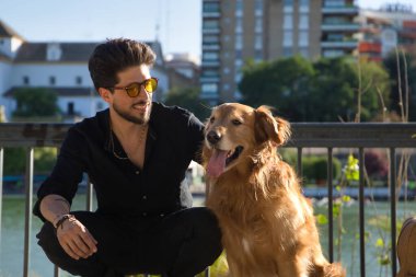 Sakallı, güneş gözlüklü ve siyah gömlekli genç bir İspanyol, köpeğiyle birlikte onu güneşin altında okşarken banka tırmandı. Hayvanlar, köpekler, aşk, evcil hayvanlar, altın.