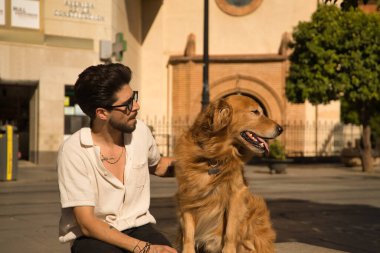Young Hispanic man with beard, sunglasses and white shirt, sitting on a bench hugging and petting his dog. Concept animals, dogs, love, pets, golden.