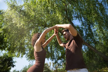 Yeşilliklerle çevrili bir parkta kolları havada dans eden çekici genç bir çift. Latin dansı konsepti, şehvetli, folklor, kentsel.