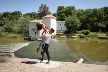Taş bir zeminde, açık hava parkında, nehrin yanında dans eden çekici genç çift. Latin dansı konsepti, şehvetli, folklorik, kentsel.