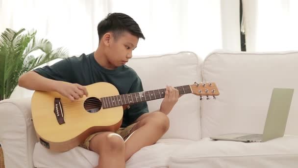 亚洲男孩住在家里 坐在白色沙发上拿着吉他 通过笔记本电脑在线学习音乐 国际音乐教育概念 网上教育 — 图库视频影像