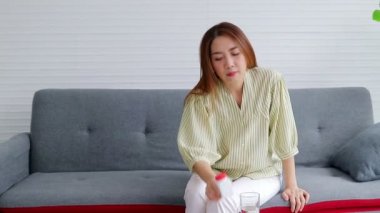 Asyalı kadının başı ağrıyor ve ateşi var. Ağrı kesici aldı ve su içti. Evdeki oturma odasında gri bir kanepede oturmak. Hastalığı evde tedavi etme kavramı. sosyal uzaklık