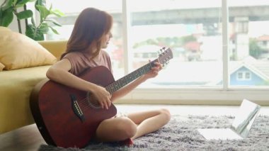 Güzel bir Asyalı kadın evinin oturma odasında gitar çalar. İnternette müzik öğrenme kavramı. Boş zaman aktiviteleri, iyi istirahatler.