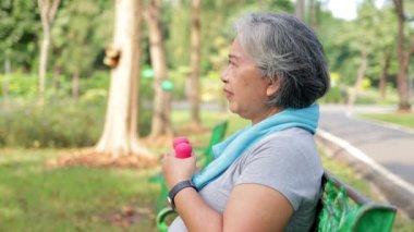 Asyalı yaşlı kadın sabah parkta oturup egzersiz yapıyor. Pembe halterleri tutar ve kollarını uzatır. fitness konsepti Yaşlıların fiziksel sağlığıyla ilgilenmek