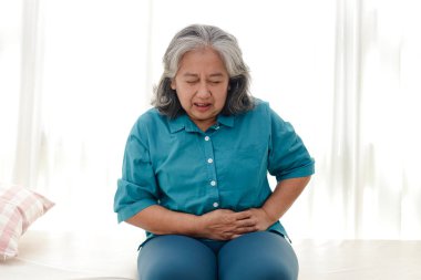 Asyalı yaşlı kadının midesi ağrıyor. O, beyaz yatağa oturdu. Yaşlılar için sağlık hizmeti kavramı. Hastalık. Sağlık sigortası.