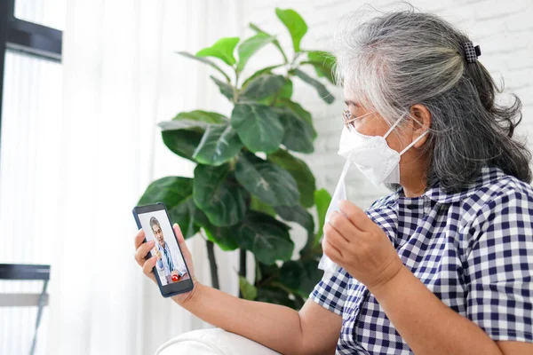 Online Medicine Concept. An elderly Asian woman has a fever. Holding a black smartphone, talking to a doctor online, asking how to use medicine. Social distancing to prevent viral infection