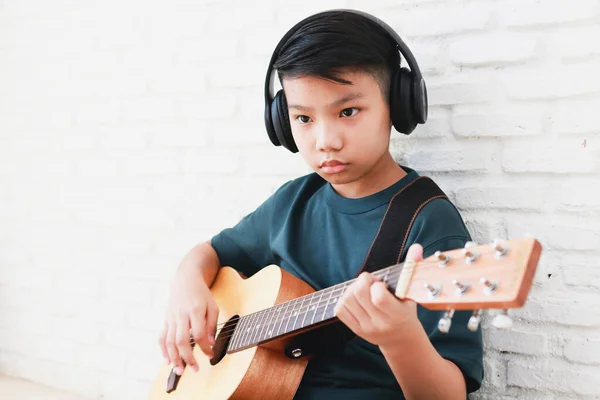 身穿黑色音乐耳机的亚洲男孩坐在屋里弹吉他 音乐学习概念 音乐熟练程度训练 — 图库照片