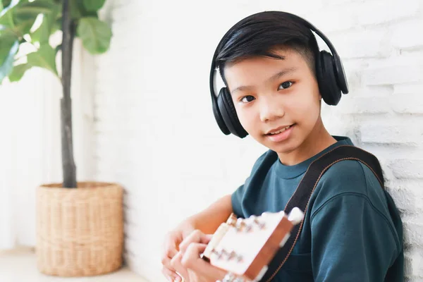 身穿黑色音乐耳机的亚洲男孩坐在屋里弹吉他 音乐学习概念 音乐熟练程度训练 — 图库照片
