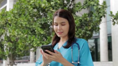 Mavi ameliyat önlüğü giyen Asyalı bir kadın doktor bir hastayla akıllı telefondan konuşuyor. Tıbbi hizmet kavramı, teknoloji ve sağlık. Çevrimiçi hizmetler.