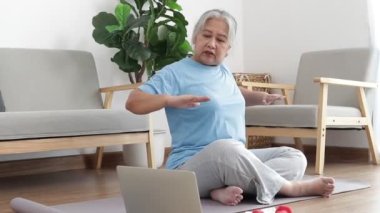 Asian elderly woman sitting at home exercising, doing exercises according to online fitness trainers. through a video call on a laptop. Social distancing, maintaining the health of the elderly