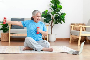 Asian elderly woman sitting at home exercising, doing exercises according to online fitness trainers. through a video call on a laptop. Social distancing, maintaining the health of the elderly