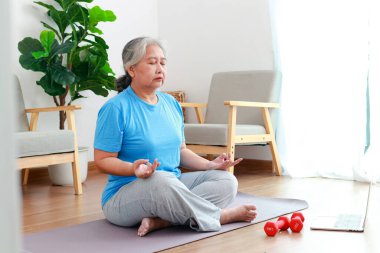 Asyalı yaşlı kadın evde egzersiz yapıyor, online fitness eğitmenlerine göre egzersiz yapıyor. Bilgisayardaki bir görüntülü aramayla. Sosyal uzaklık. Yoga yaparken meditasyon yap