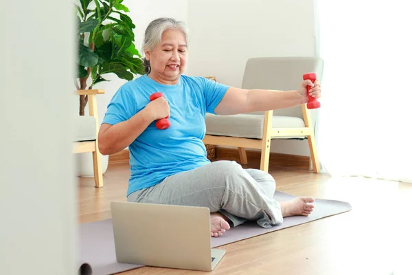 Asian elderly woman sitting at home exercising, doing exercises according to online fitness trainers. through a video call on a laptop. Social distancing, maintaining the health of the elderly