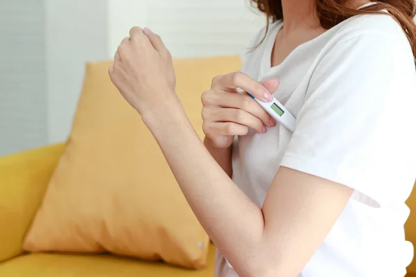 Asian woman sick with flu treating herself at home Hold the digital thermometer in your armpit to check your body temperature. concept of health care
