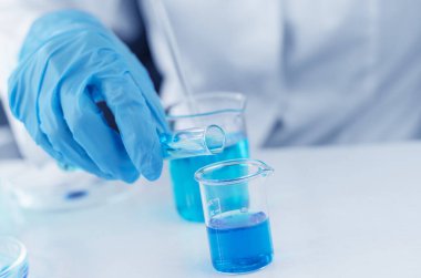 Beyaz önlüklü bir bilim adamı deney tüpünden laboratuvar bardağına sıvı döküyor. Aşı araştırma ve geliştirme