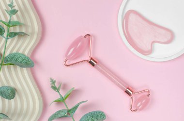 Pembe arka planda, geometrik şekilli yüz masajcısı. Pembe arka planda yeşil yapraklı Gua Sha yüz masajı araçları