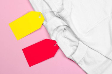 Pembe arka planda iki beyaz tişört ve boş fiyat etiketleri. Kıyafetlerde boş etiket