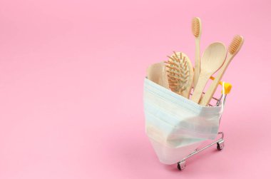 İnternetten ekolojik ürünler satın alma kavramı. Pembe arka planda koruyucu bir tıbbi maskeyle alışveriş arabasında ahşap şahsi bakım ürünleri.