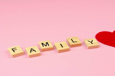 Aile kelimesi ahşap harflerden ve pembe arka planda yarım bir kırmızı kalpten oluşur. Aile Günü.
