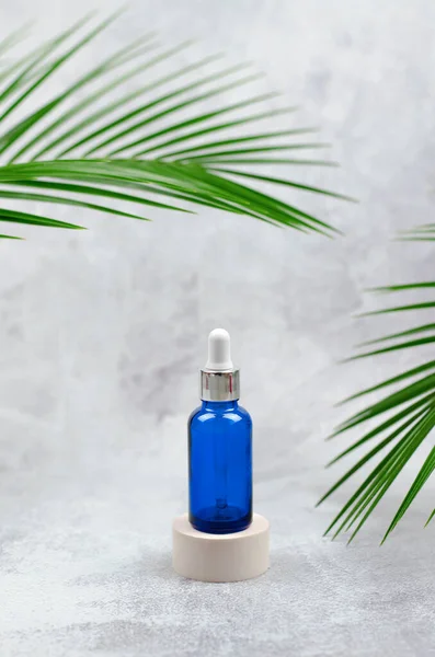 A blue cosmetic bottle with a pipette on a round beige podium on a light gray background with palm leaves. Natural care cosmetics for face and body.