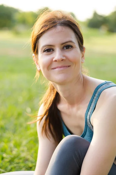 Genç bir kadın parkta yoga minderinde oturuyor. Güzel genç sporcu kadın eğitimden sonra dinleniyor..