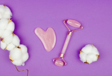 Gül kuvars gua sha masaj kazıyıcısı ve mor arka planda doğal pamuklu yüz masajı silindiri. Yaşlanma karşıtı tedaviler. Yüz bakımı