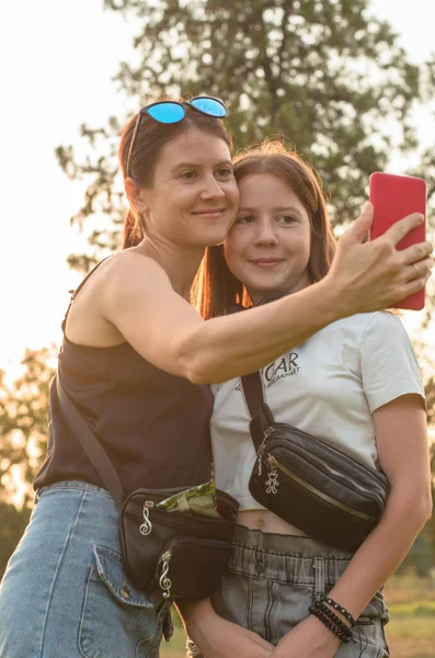 Mom and daughter take a selfie in nature. Family portrait. Family walk in the park. Happy mom and teenage girl are photographed on a summer evening.