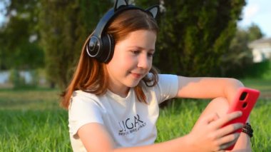 A teenage girl sits in a park on the grass with a smartphone in her hands. 12 year old girl listening to music in headphones sitting in the park on the grass
