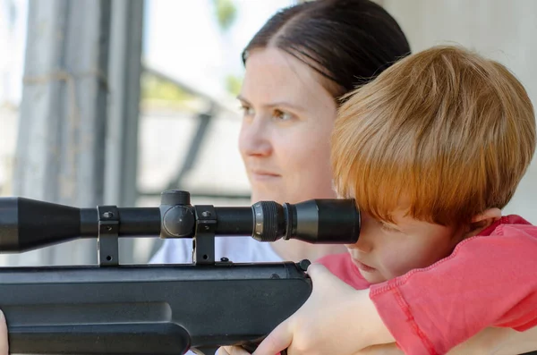 A Ukrainian woman teaches her son how to shoot a rifle. A 5-year-old boy looks through the optical sight of a sniper rifle. Child with a gun.