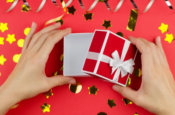 Open gift box in female hands on a red festive background, close-up. Woman opening a small gift box