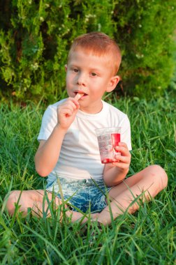 Beyaz tişörtlü bir çocuk doğada dondurma yer. Küçük kızıl saçlı çocuk parkta çimlerin üzerinde oturarak dondurma yiyor. Mutlu çocukluk.