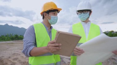 Asyalı inşaat işçileri güvenlik yeleği, güvenlik kaskı, koruyucu yüz maskesi giyer. Salgın sırasında inşaat alanında birlikte çalışırlar. Tablet taslakları, hastalık önleme kağıtları, proje üzerinde birlikte çalışan çeşitli uzmanlar tutarlar.