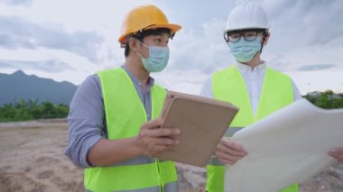 Asyalı inşaat işçileri sıhhiye maskesi ve tam güvenlik üniforması takıyorlar. Salgın sırasında inşaat alanında birlikte çalışıyorlar. Tablet tutuyorlar. 