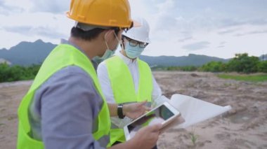 Asyalı erkek inşaat işçisi inşaat projesinde güvenlik yeleği ve sert kask takıyor, takım arkadaşı, dijital tablet planları kağıt, mühendis ve mimar birlikte çalışan çeşitli uzmanlar bir proje üzerinde birlikte çalışıyorlar.