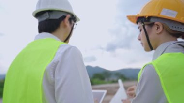 Düşük açılı çekimde, yüksek açılı çalışan iki çalışan, çalışan proje üzerinde tartışırken, doğal dağ sırasının arka planında ve berrak gökyüzünde, bir inşaat inşaatçısı ekibi, çevre dostu