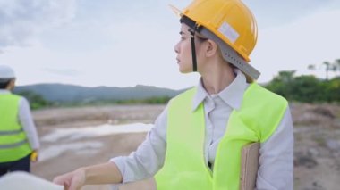 Genç bayan mühendis, mimari taslak çizimlerine, yapı planlamacısına, emlakçıya, önce güvenlik, açık hava inşaat sahasına, imalat projesi çalışanına, proje üzerinde birlikte çalışan çeşitli uzmanlara bakarak güvenlik vitesi takıyor.