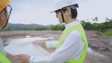 Çamurlu doğal inşaat sahasında dururken inşaat planlarını tartışan aktif bir müteahhit ekibi, havada bir yapı çizmek için el yapımı planlar, çevre dostu inşaat projesi, proje üzerinde birlikte çalışan çeşitli uzmanlar