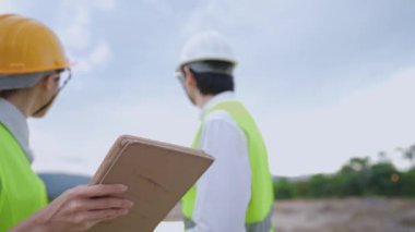 Arka görüş, toprak inşaat sahasında çalışan kendini adamış uzman teknisyenler, genç bayan asistan projenin detayları, işyeri çeşitliliği ve takım çalışması konsepti hakkında bilgi toplamak için dijital tablet kullanıyor