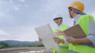 Asyalı genç mühendis miğferi, inşaat sahasındaki çalışma projeleri, uzmanlık alanı, emlak yatırımları, iş vites tasarım planları, proje üzerinde birlikte çalışan çeşitli uzmanlar, güvenlik yeleği miğferi giyiyor.