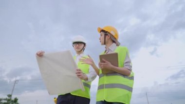 İnşaat alanının dışında duran güvenlik ekipmanı giyen Asyalı işçiler, yapı projesi üzerinde inceleme ve tasarım, plan çizim kağıdı, takım çalışması koordinatörü, proje üzerinde birlikte çalışan çeşitli uzmanlar