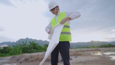 Asyalı erkek mühendis mimarisi güvenlik yeleği giyer. Kask katlama planı çizer. İnceleme işini bitirdikten sonra, uzmanlık alanı, zaman içinde çok çalışan çalışanlar, açık alan, inşaat projesi üzerine profesyonel tasarım. 