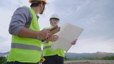 Asyalı mimari mühendisleri güvenlik üniforması giyer, inşaat alanındaki inceleme sırasında kendilerini tehlikeden korurlar, tablet taslak kağıtlarını tutarlar, güneşli günlerde çok çalışırlar, çeşitli uzmanlar proje üzerinde birlikte çalışırlar.