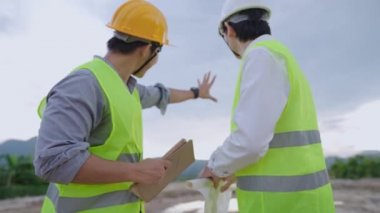 Asyalı mimari ve mühendis işçiler inşaat sahasında birlikte çalışan güvenlik yeleği ve güvenlik kaskı giyerler, inşaat alanına bakan tablet taslak kağıtları, takım çalışması planlama, proje üzerinde birlikte çalışan çeşitli uzmanlar