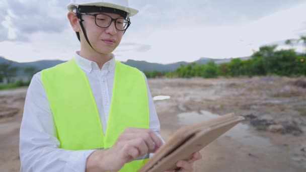 アジアのエンジニアの労働者は 屋外の建設現場 専門の作業現場に立って 建築設計図 分析のアイデアと計画の進捗状況 土地上のプロの設計建築プロジェクトを見て 眼鏡と保護作業の摩耗を着用します — ストック動画