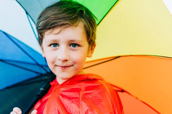 Retrato de um menino da escola com guarda-chuva de arco-íris atrás. O miúdo tem um guarda-chuva colorido no ombro. Criança alegre em uma capa de chuva vermelha segurando guarda-chuva multicolorido — Fotografia de Stock