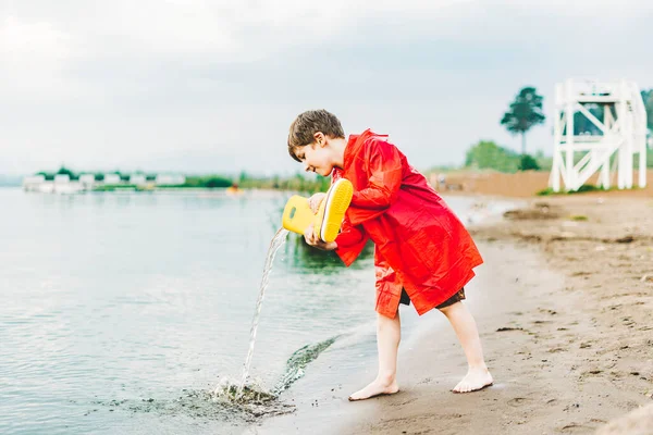 Junge in rotem Regenmantel schüttet Wasser aus gelben Gummistiefeln in den See. Kind spielt mit Wasser am Teich. Kind vergnügt sich im Freien am Flussufer — Stockfoto