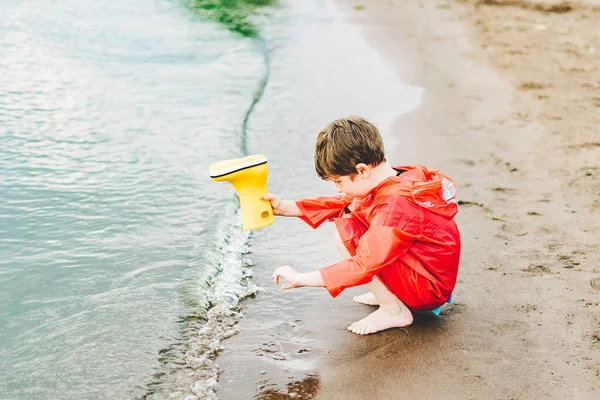 Niño en un impermeable rojo vierte agua de bota de goma amarilla en el lago. Niño jugando con agua en el estanque. Niño divirtiéndose al aire libre cerca de la orilla del río —  Fotos de Stock