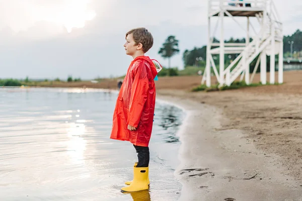 Niño con un impermeable rojo y botas de goma amarillas jugando con agua en la playa. Niño de la escuela en un abrigo impermeable saltando en el agua en el mar. Niño divirtiéndose con olas en la orilla —  Fotos de Stock