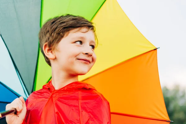Retrato de um menino da escola rindo com guarda-chuva arco-íris atrás. O miúdo sorridente tem um guarda-chuva colorido no ombro. Criança alegre em uma capa de chuva vermelha segurando guarda-chuva multicolorido — Fotografia de Stock