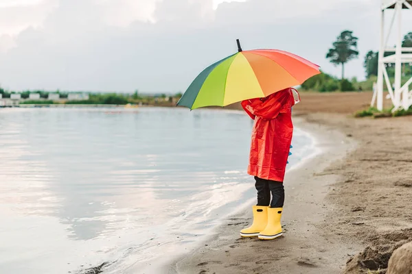 Boy in a red raincoat and yellow rubber boots stands at river bank and holding rainbow umbrella. School kid standing still near autumn lake. Child wearing waterproof clothes at shoreside